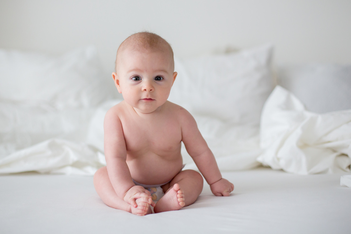 Cute little baby boy in diaper, smiling at camera in white bedroom, cute toddler sitting on bed, smiling