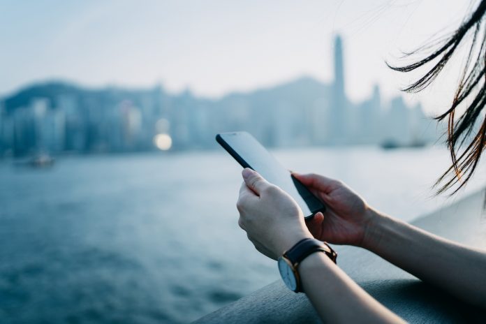Close up of young woman using mobile phone by the promenade of Victoria Harbor with urban city skyline in background