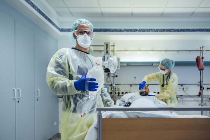 Photo of doctor in emergency care unit of a hospital holding bag valve mask and standing next to a COVID-19 patient in a bed being attended to by another healthcare worker