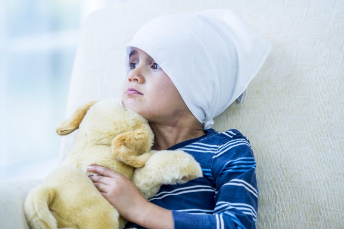 Young, male, brain cancer patient wearing a hat/head covering is sitting in bed With a Toy Dog.
