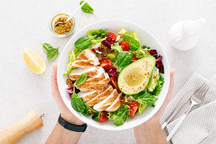 Grilled chicken meat and fresh vegetable salad of tomato, avocado, lettuce and spinach. Healthy and detox food concept. Ketogenic diet. Buddha bowl in hands on white background, top view