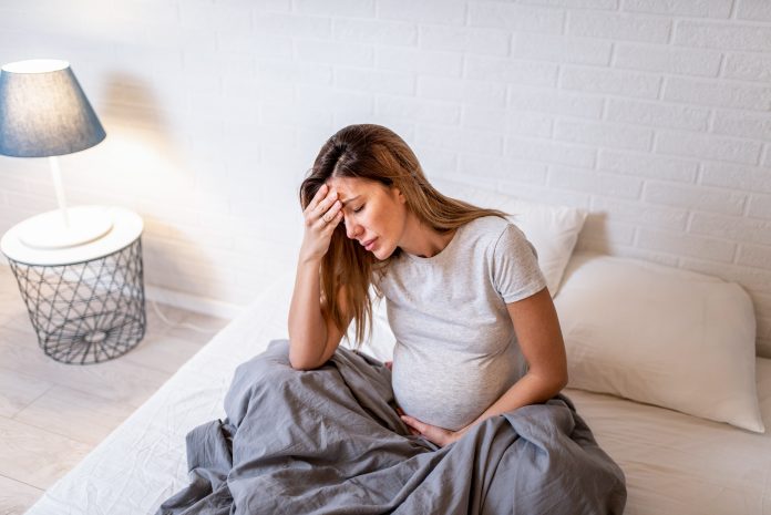 Young pregnant woman sitting on floor and holding her head as she is experiencing a migraine.