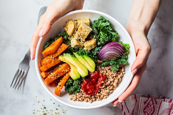 Woman hands eating vegan salad of baked vegetables, avocado, tofu and buckwheat buddha bowl, top view. Plant based food concept.