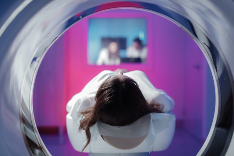 Image of a young female patient lying in a CT machine waiting to have a scan, such as photon-counting CT. Two healthcare professionals can be seen monitoring the scan through a window in the background of the image.