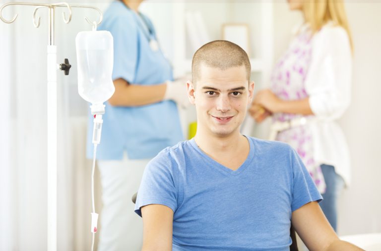 Photo of an young man receiving chemotherapy at home. In background his girlfriend talking with nurse. Illustrating cancer epigenetics.