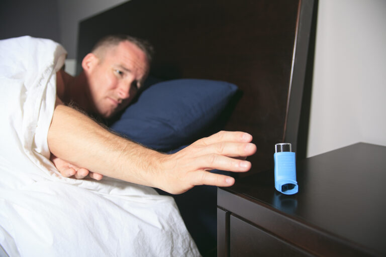 A white man lying in bed before going to sleep reaching for a blue asthma inhaler on the bedside table.
