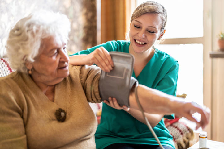 Young female carer measuring senior woman's blood pressure at home to assess health risks of various conditions including dementia