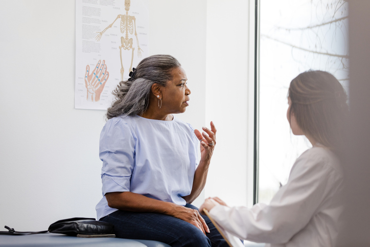 Confused older, Black woman looks out window while talking to female doctor facing patient to illustrate that people with different genetic ancestry may have different prevalence of brain disorders.
